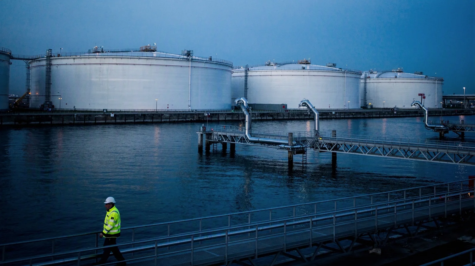Coastal petroleum products terminal at blue hour, storage tanks and loading arms with no ship at berth, calm water reflecting cold sky