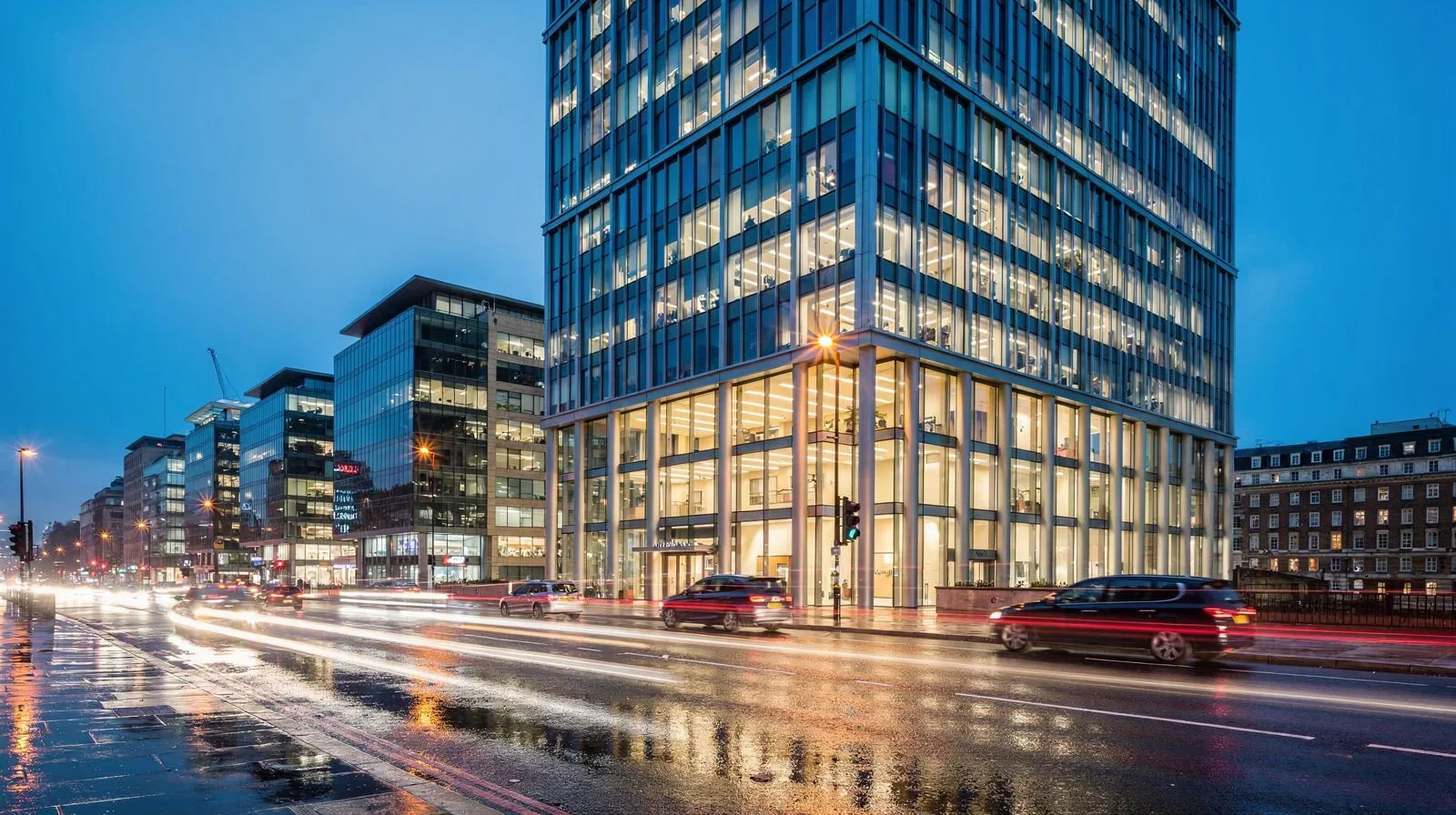 A discreet London private-wealth office at blue hour, with a warmly lit meeting room behind glass and Mayfair traffic blurred below