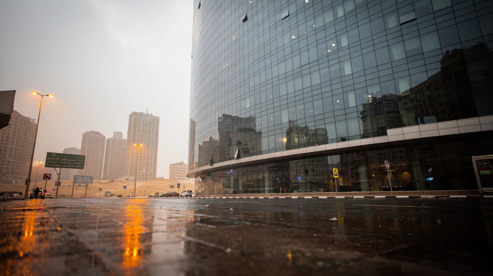 A close view of Cairo’s contemporary glass towers after rain, with luminous street reflections and a single exchange-like facade receding into soft desert haze