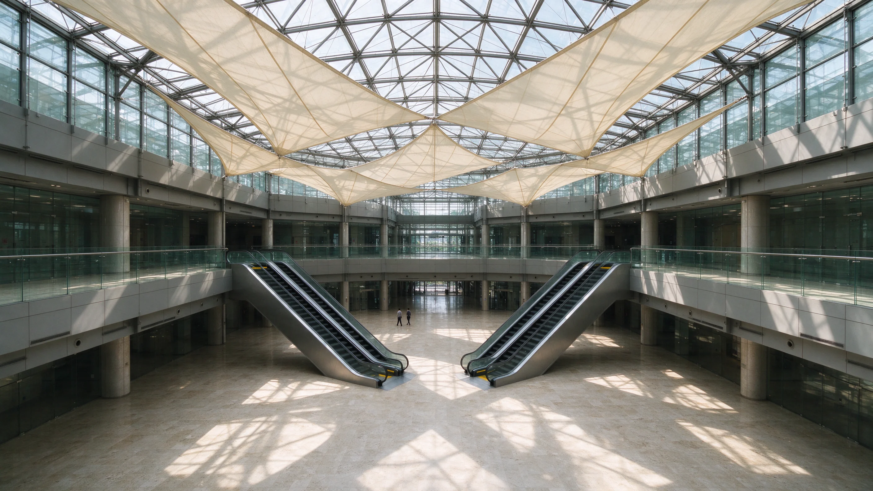High angle across a Bengaluru tech-park atrium at bright midday, sparse foot traffic on polished floors beneath blank fabric sails, escalators mostly empty
