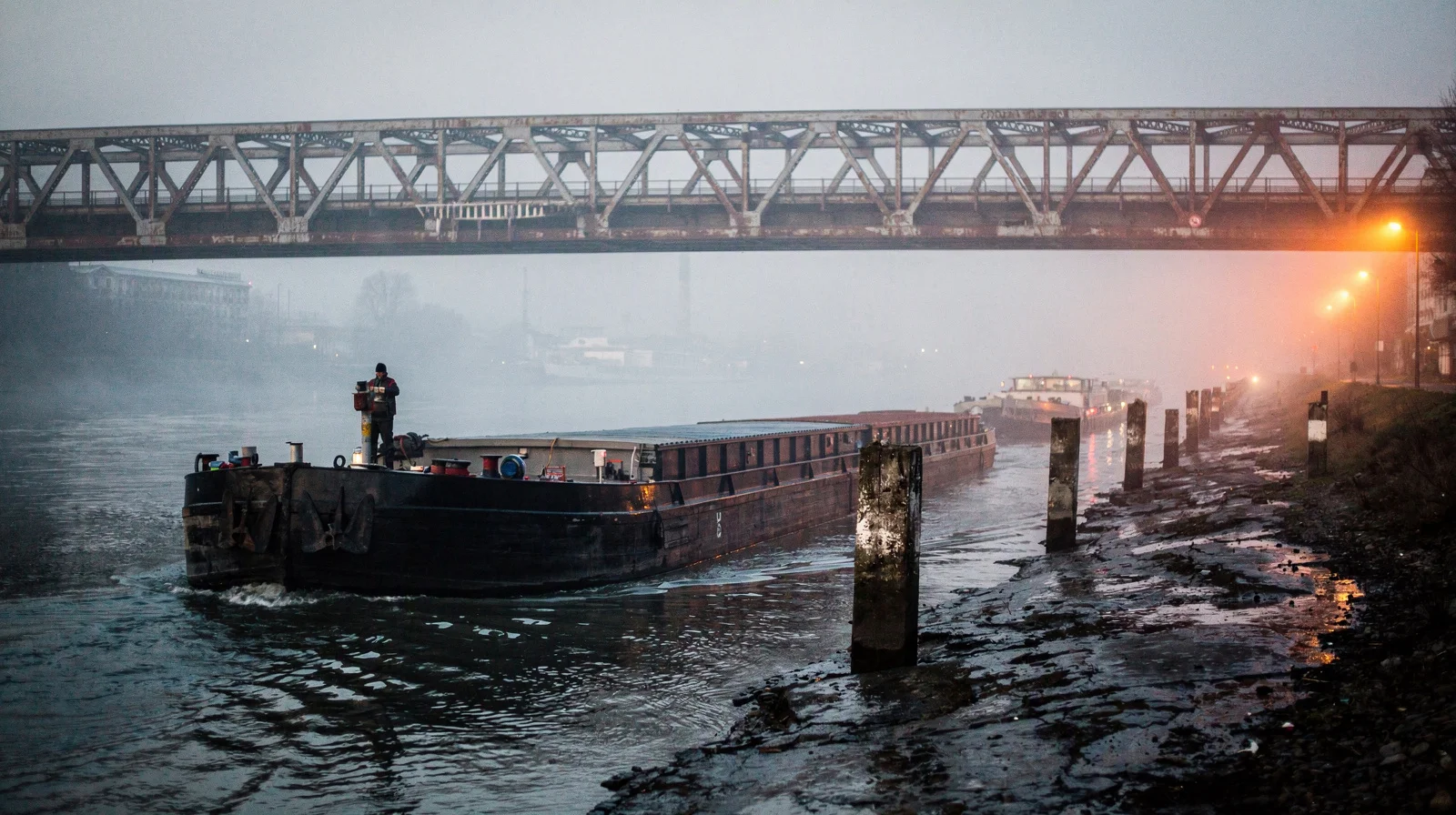 Fog-bound Rhine cargo barges under a steel bridge at dawn, low water and sodium lights on the bank