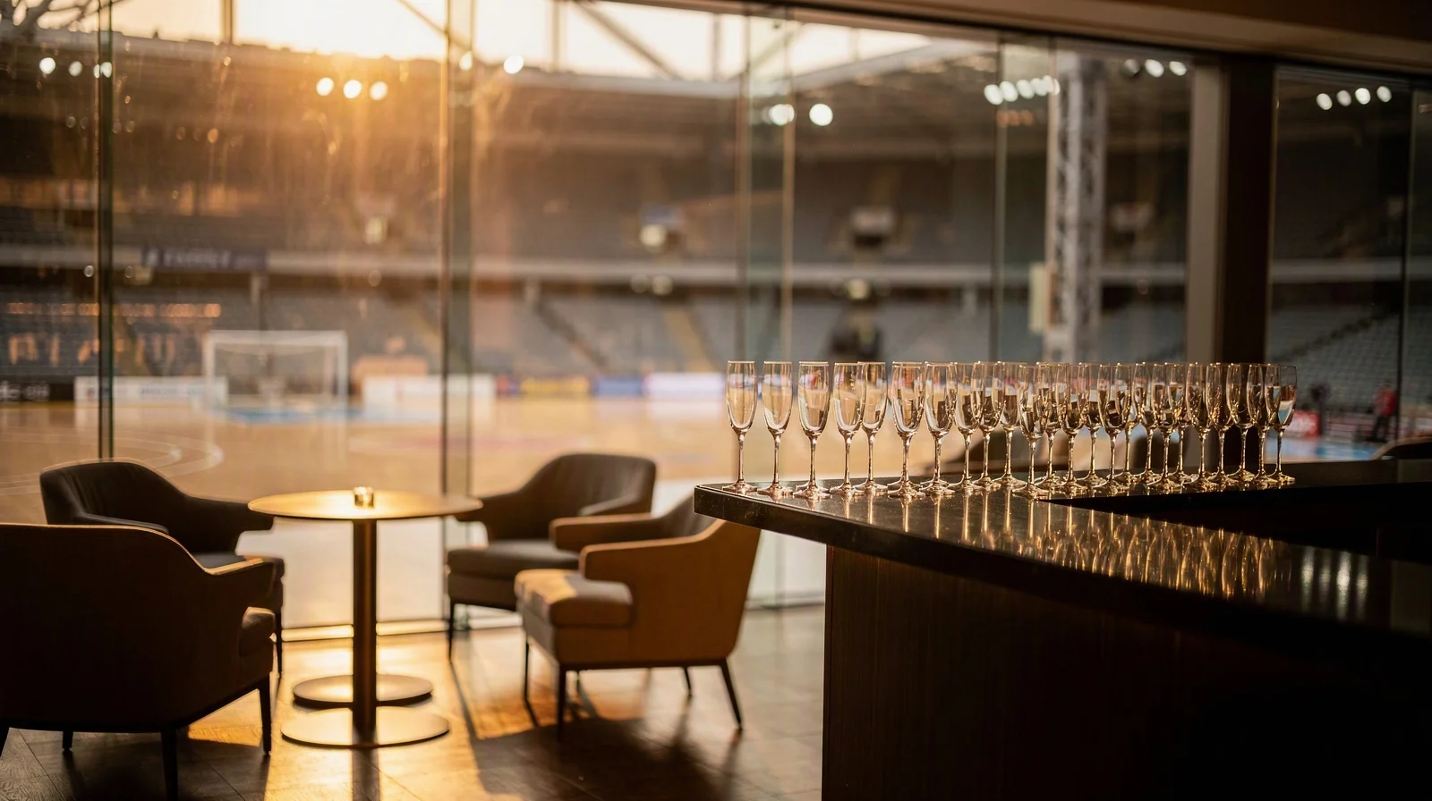 Empty premium stadium hospitality suite with champagne flutes and floor-to-court glass, warm evening light