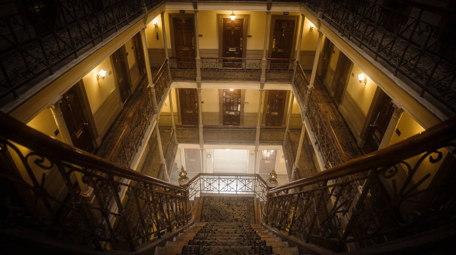 Grand London mansion-block interior atrium with multiple apartment doors on each landing, warm sconce light and patterned carpet runner