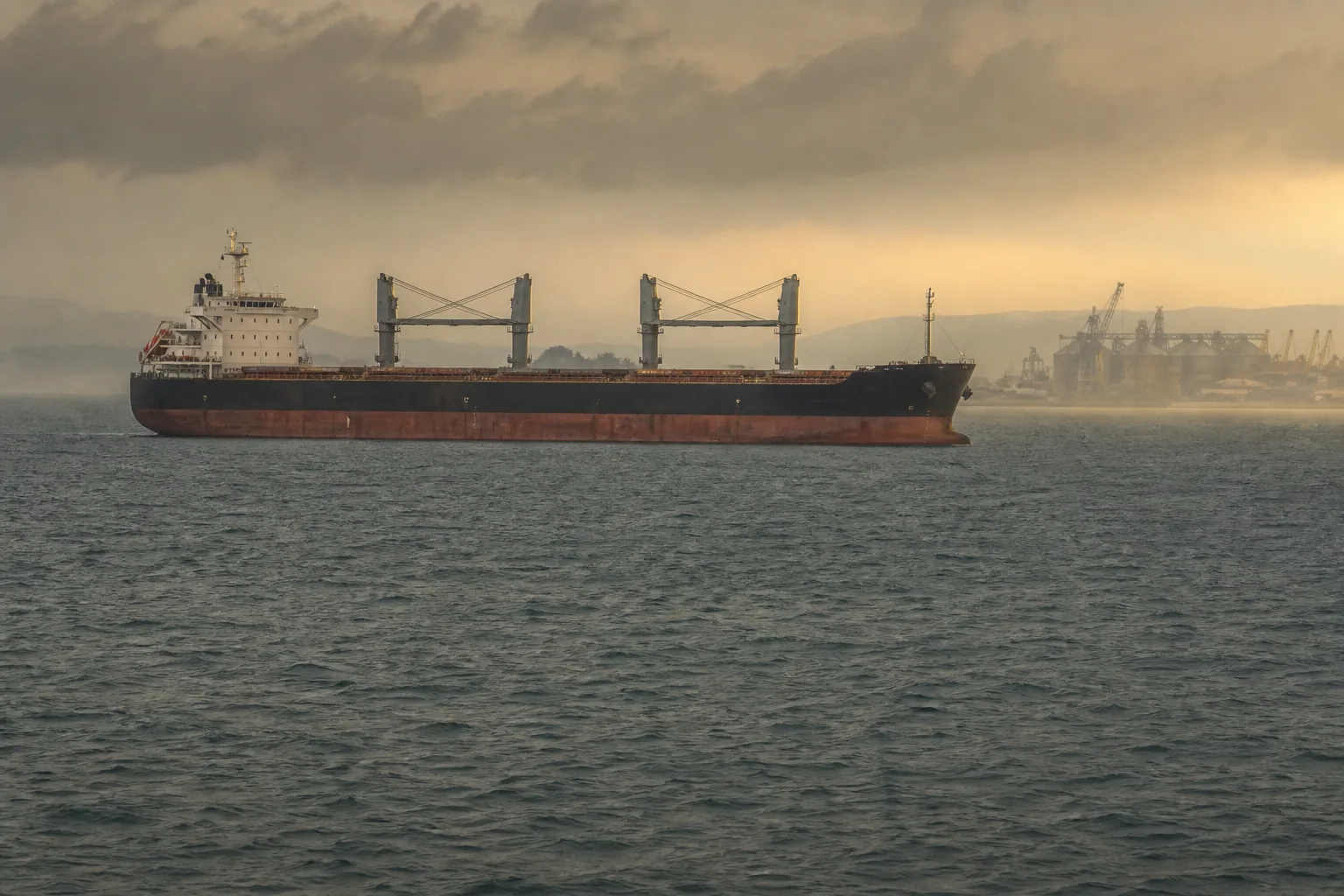 A laden bulk grain carrier waiting offshore in grey Black Sea water, idle silos and loading arms hazy on the distant coast