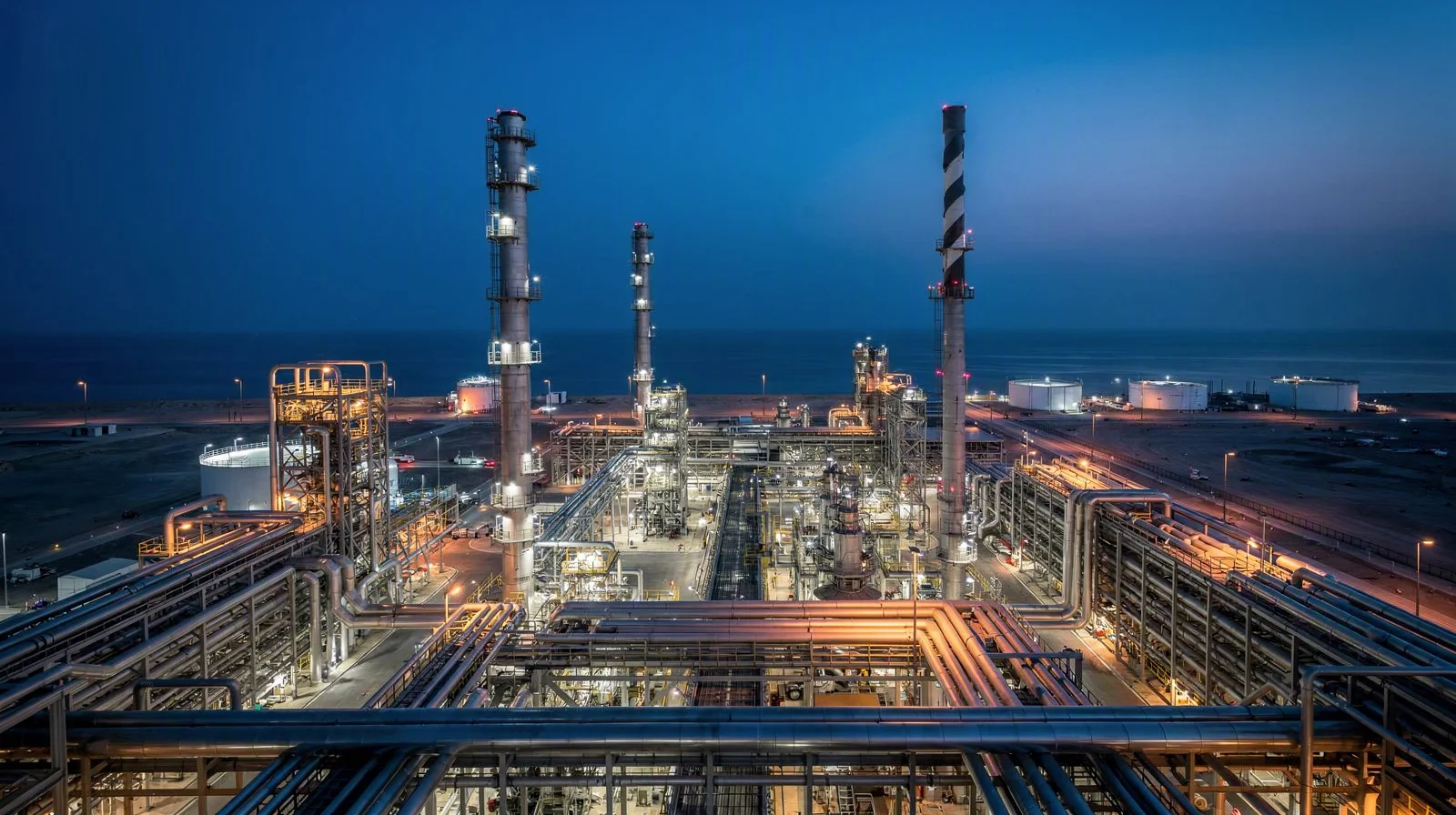 Oman’s coastal refinery and chemical-processing infrastructure at blue hour, with luminous pipes and towers beside a dark Arabian Sea horizon