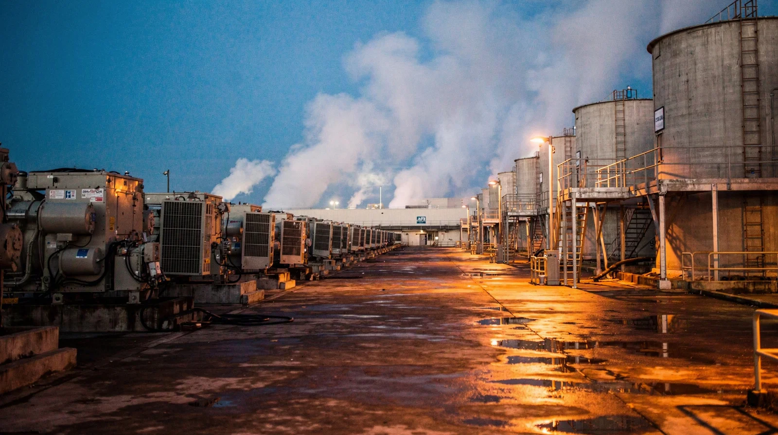 Hyperscale data center cooling towers and backup generators at dusk, steam rising under sodium lights on wet concrete