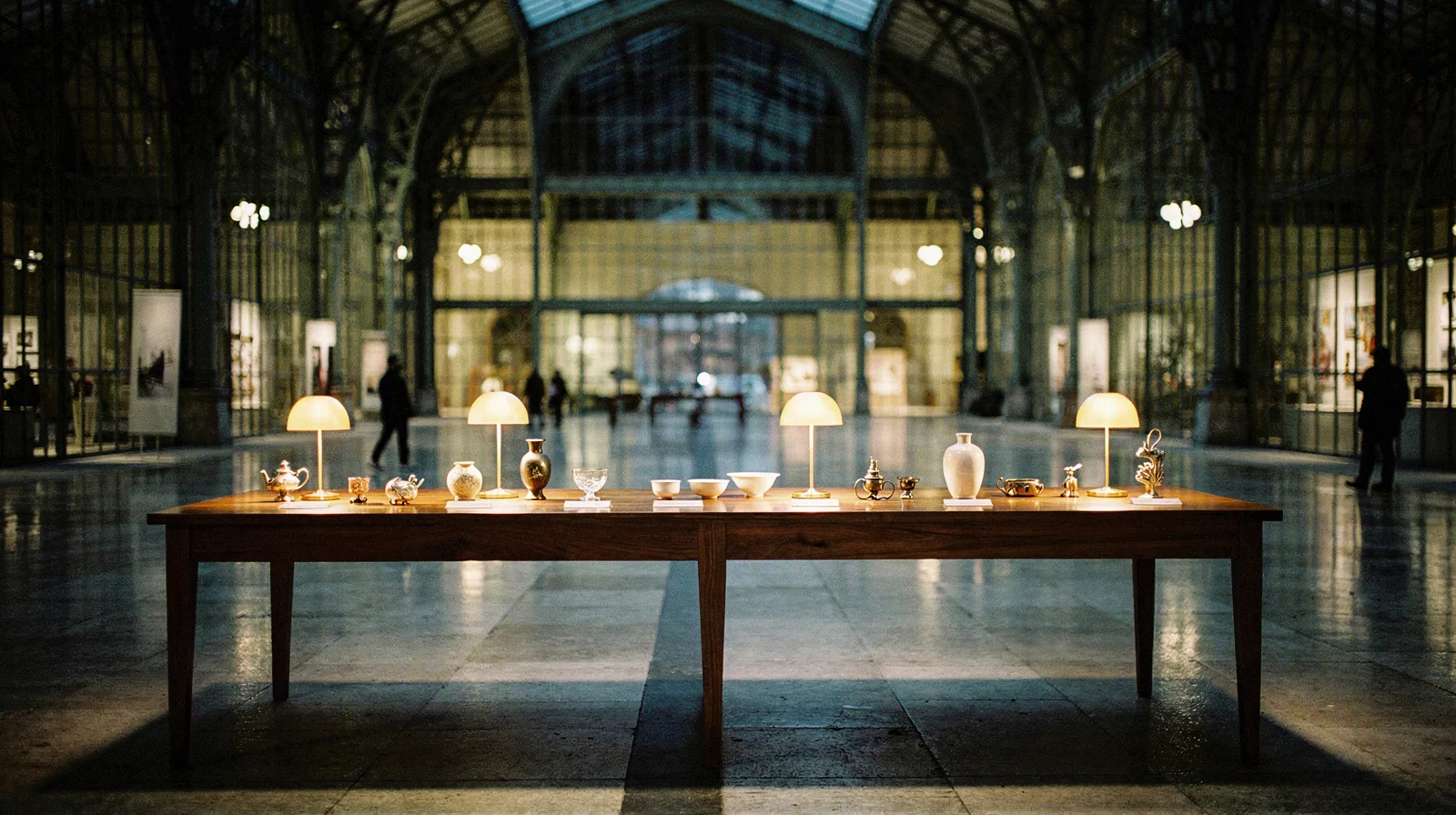 Illuminated craft objects arranged on a walnut display table beneath a grand glass-and-steel exhibition hall