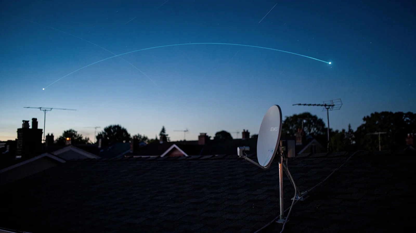 Suburban rooftop at dusk with a small white satellite dish against a sky streaked with faint satellite trails and a thin laser-like line between two points of light